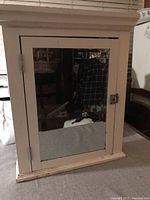 Front view of the white heavy medicine cabinet showing a rectangular mirror on the door, metal hinges on left, and a broken lock mechanism on the right side of the door.