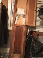 Wooden wall-mounted ironing board in vertical folded position on a wall, showing decorative fabric padded cover at top.