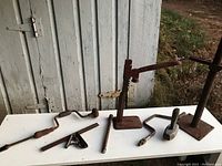 Photo showing vintage mechanical jack, two hand crank drills, metal pipe, and square-shaped tool on a white table outdoors. Items aged with rust and wear visible.