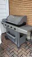 Front view of the grill showing four control knobs, lower storage shelf, and wheels.