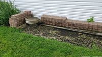 View of the stacked rectangular bricks alongside two round stone pathway pavers placed on wooden pallets by a white siding wall.