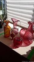 Variety of six cranberry glass items displayed on a windowsill with natural light showing colors and textures.