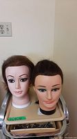 Front view of two mannequin heads with painted facial features and synthetic hair, placed on a shelf.