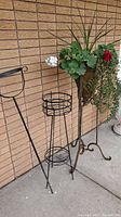 Metal plant stand, garden plant stand and conical planter in a stand placed outside against a brick wall with visible plants inside the conical planter.