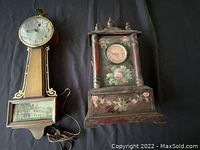 View of two antique decorative wall clocks: one with large house image on wooden panel and round clock face; second with floral painted wooden case and small round clock face, both lying on fabric background.