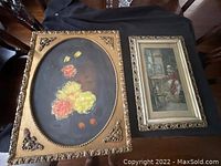 Photo showing two antique picture frames on a black cloth surface, one oval with colorful flowers painting, the other rectangular with domestic interior scene painting.