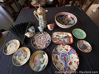 Overview of all items in the lot including pitcher, large bowl, multiple plates, cups and folding fan laid out on black cloth.