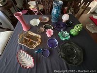 Photo showing a collection of colorful glass and ceramic decorative items laid out on a table including vases, plates, and small ornamental dishes