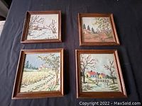 Four framed embroidery artworks displayed on a dark fabric background. Each piece features a different house and tree countryside scene.