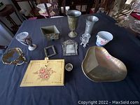 Table with various metal and glass decorative items and kitchenware displayed on a black cloth, including a floral painted metal book box, photo frames, goblet, candy bucket, heart-shaped dish, and glass with metal stand.