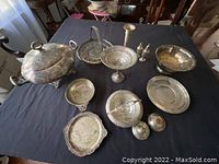 Overview of 11 assorted mixed metal serving and tableware items on a table including covered bowl, trays, compote dish, ladle, salt & pepper shakers, candlestick, and bowls.