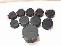 Front view of 11 riflescope lens caps arranged with brand embossing visible including Millett and Leupold logos.