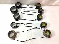 Lot of 8 riflescope lens caps in various sizes and models with clear and yellow transparent covers connected by elastic cords, photographed against white background.