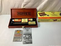 Open solid wood case with Hoppe's gun cleaning kit items inside including cleaning lotion, lubricant oil, rod ends, rod pieces, cleaning patches, and cloths, with original box in the background.
