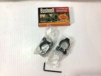 Complete lot showing Bushnell box, two 1-inch scope mount rings sealed in plastic, and included hex wrench on a white background.