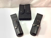 Black Cordura double magazine case with double row pouch, secured by two snap-button flaps, positioned centrally above two black leather belt holsters on a white background.