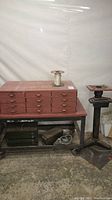 Photo shows a large red multi-drawer storage cabinet on a metal bench with diverse woodworking items, spool of wire on top, and the bench grinder base stand next to it.