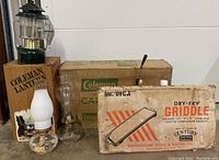 Lot contents showing Coleman lantern on wooden box, two oil lamps with chimneys, and original boxed camp stove and griddle.