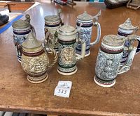 Set of seven earthenware collectible steins with metal lids on wooden surface, showing varied designs including ship, train, hunting, and wildlife themes.