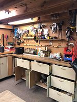 Wide view of garage workspace featuring wall-mounted pegboard with various hanging tools, open drawers and cabinets containing supplies and tools.