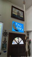 Wide shot of wall showing framed ship painting above door, wooden ship wheel, anchor, seaside scene, and coiled ropes.