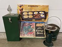 Photo showing Coleman two burner camp stove, green metal carrying case, Presto electric griddle in box, and a metal bracket.
