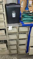 Pen and Gear paper shredder on top of rusted metal organizer with drawers. Shredder shows paper inside window.