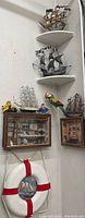 Corner wall display of nautical decor showing three model ships on corner shelves and two shadow boxes, one life ring and two parrot figurines around them.