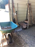 Wide view of all items including wheelbarrow, ladder, shovels, pitch forks, and wood pieces leaning against a wall.