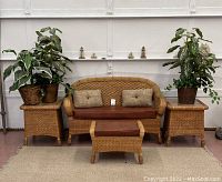 Front view of wicker bench with brown cushion and two smaller cushions flanked by two wicker end tables with three potted artificial plants on and beside them.