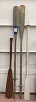 Four wooden decorative oars of various lengths and colors leaning against a wall, showing overall condition and size difference.