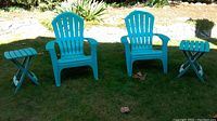 Two teal resin Adirondack chairs and two matching folding side tables on grass outdoors.