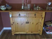 Front view showing wooden buffet cabinet with two drawers, two doors, and a top shelf with various glassware on it.