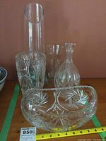 Photo showing crystal and glass vases along with crystal decanter and bowl arranged on a surface against a peach wall, includes measuring tape and lot label.