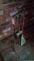 Photo showing metal fireplace accessory set — a stand with three hanging tools including a poker, brush, and shovel, positioned on a brick hearth.
