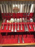 Front view of flatware set showing knives, forks, large spoons, small spoons, and a decorative pierced serving spoon in a red velvet box.