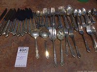 Full view of the flatware set on a rusty surface, showing all pieces including knives, forks, spoons, and specialized serving utensils.