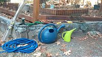Blue expandable garden hose coiled on ground, large blue plastic watering can, small green watering can, wire plant support stakes, and hand tools on stone ledge.