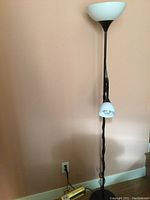 Black floor lamp showing the height, white plastic shade at top, smaller shade mid-stand, and cord on wooden floor with a peach colored wall and electrical outlet behind.