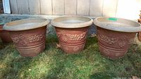 Three reddish-brown plastic planters with beige rims and floral/garland design, placed on grass outdoors, showing front view.