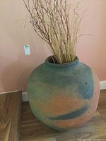 Full view of the large rounded pottery container with faux grass and twigs inside, showing the matte textured surface in blended blue and orange tones against a wooden floor and peach wall background.
