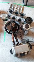 Photo showing metal muffin trays, ring molds, cast iron pans, electric waffle maker, kettle grouped together on floor