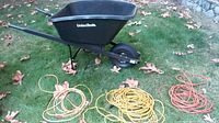 Full view of black plastic Union Tools wheelbarrow on grass with three coiled extension cords (orange, yellow, and red) beside it.