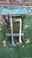 Front view of folding workbench with splitting maul and axe resting against it on grass with scattered leaves.