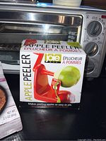 Photo showing boxed Joie apple peeler and reflective toaster oven behind