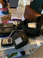 Three cases and two cameras on a wicker bench, including the black Bushnell camera case, black Handycam case, and silver Sony Cyber-shot camera