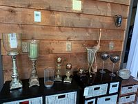Wide shot showing all items on black cabinet against wooden wall, including hurricane candleholders, mosaic glass goblets, smaller candleholders, wire holder with wooden ball, and wooden bird hanging.