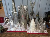 Overview of multiple decorative Christmas trees and figurines including sparkly silver cone trees, glass angels, and a white layered glitter tree displayed on fabric with red snowflake print.