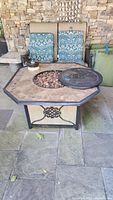 Full view of octagonal propane fire pit with tiled top, blue patterned cushions on chairs behind, cover partially off revealing lava rocks inside.