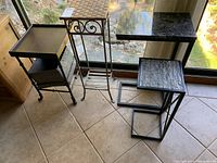 Lot viewed includes four items: metal plant stand with wicker top, metal plant stand with wooden top and scrollwork, snack table with stone tile top, snack table with laminate top.